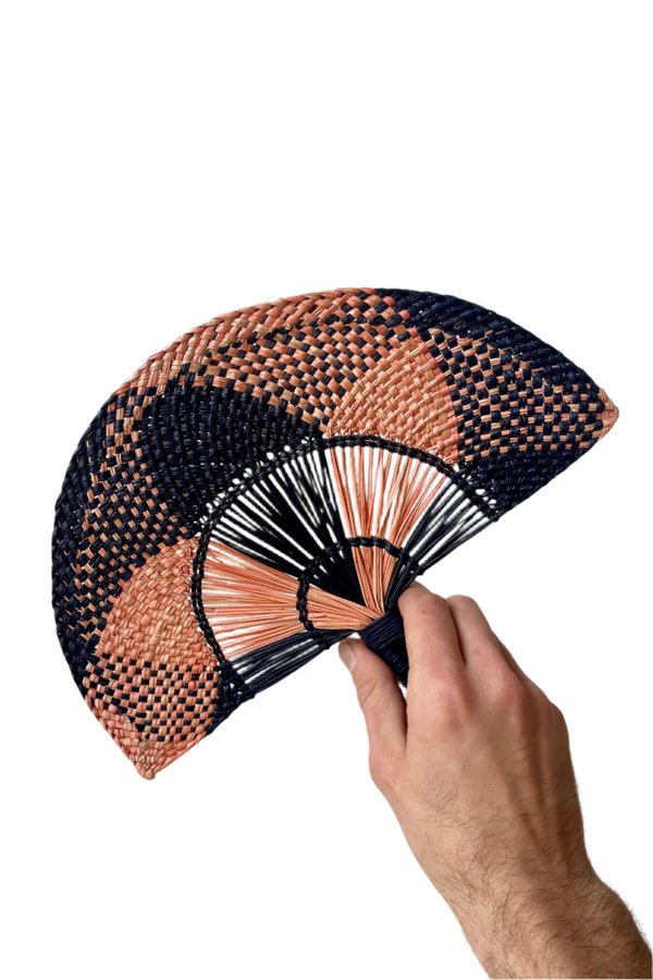 Toquilla Flat Hand Fan Navy & Pink of palm fiber and wire. A hand holds it in front of a white wall.