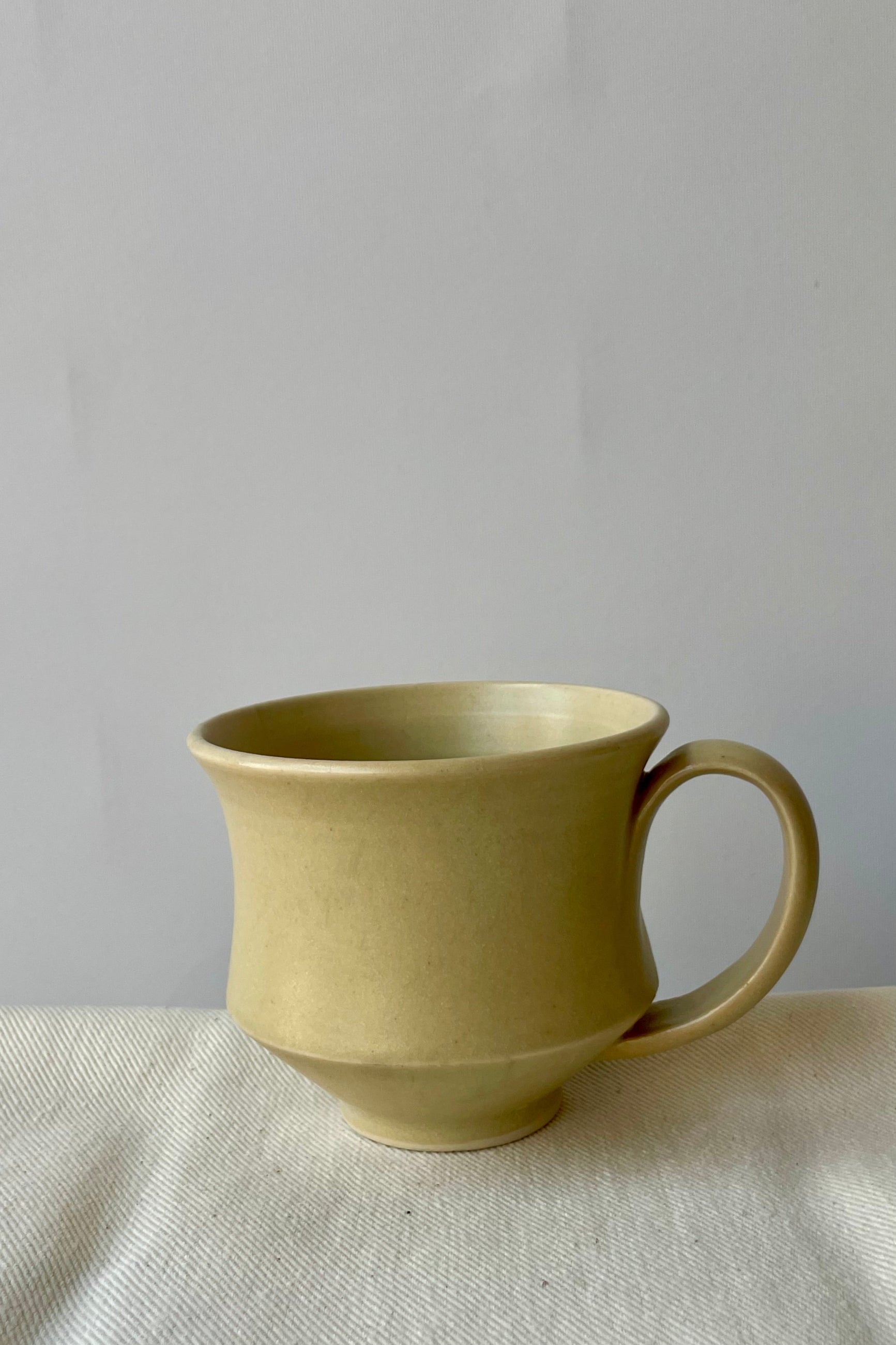 Photo of Little Fire Ceramics hourglass mug with butter glaze. Shown from the side. The mug sits on a white glass against a white wall. ©Sprout Home