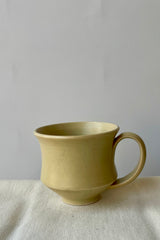 Photo of Little Fire Ceramics hourglass mug with butter glaze. Shown from the side. The mug sits on a white glass against a white wall.