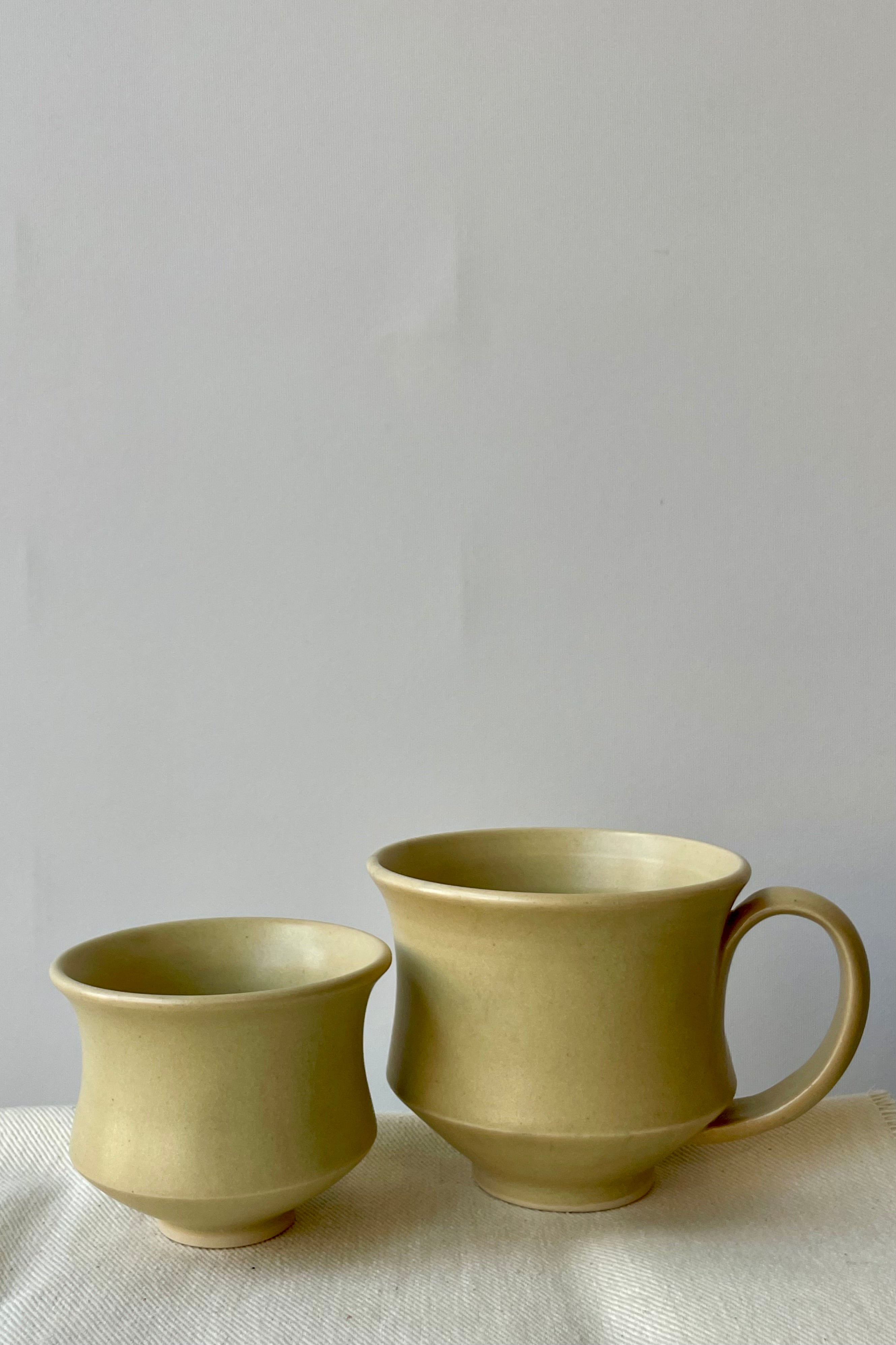 Photo of Little Fire Ceramics hourglass mug and cup with butter glaze. Shown 
side by side. Cup and Mug on a white glass against a white wall