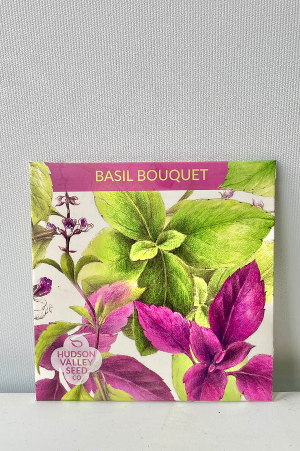 A colorful purple and green seed packet. This seed packet is called Basil Bouquet, is square in shape and features drawing of basil plants leaves and flowers. Seed packet is photographed against a white wall.