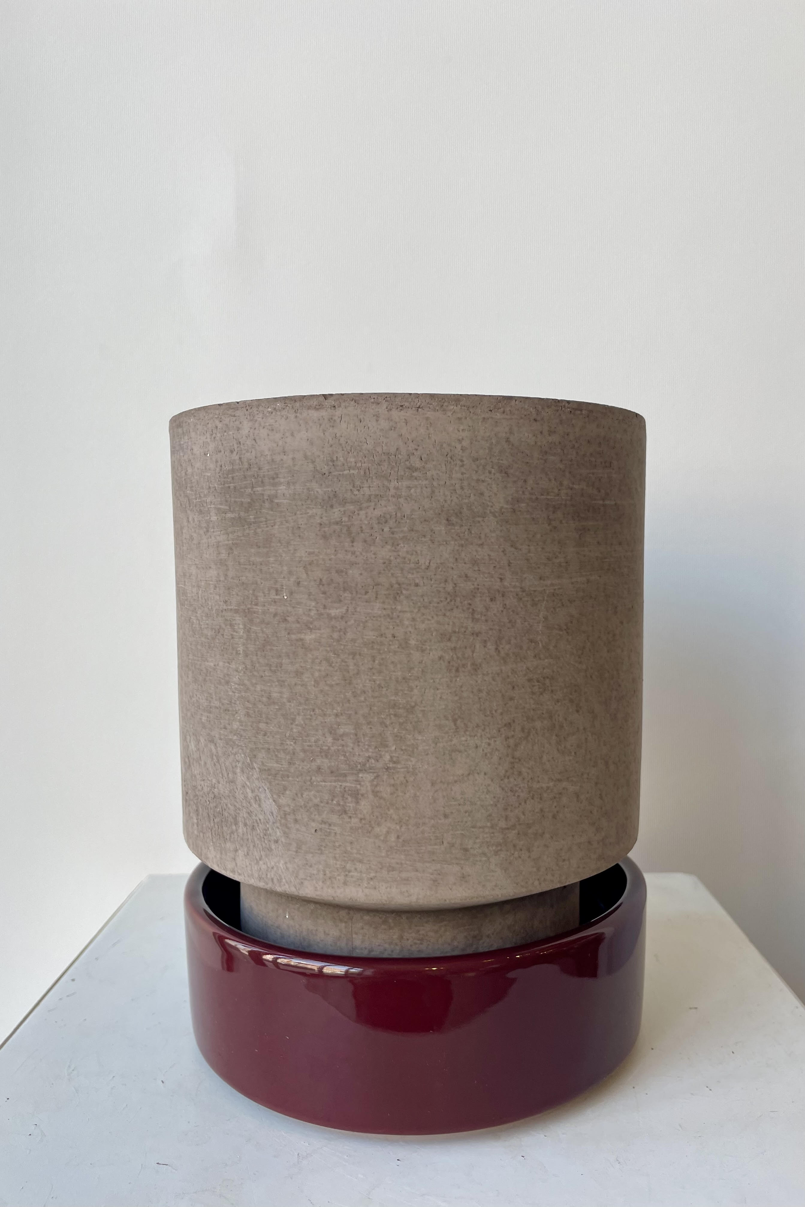 Photo of a Hoff Pot and saucer on a white table in a white room. The pot is gray and sits within a deep burgundy glazed saucer.