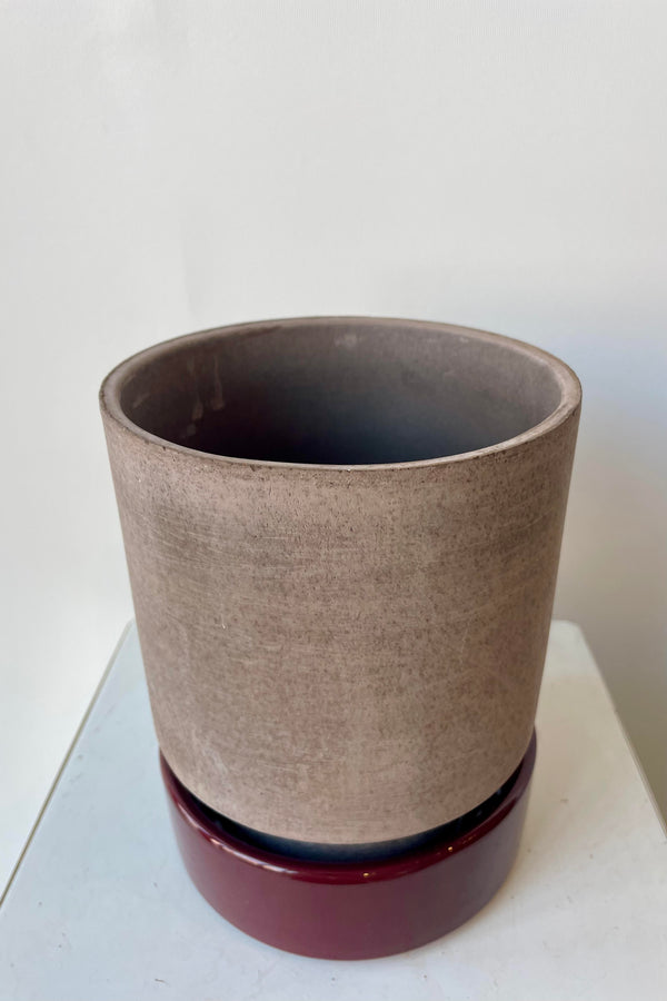 Photo from above of a Hoff Pot and saucer on a white table in a white room. The pot is gray and sits within a deep burgundy glazed saucer.