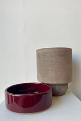 Photo of a Hoff Pot and saucer on a white table in a white room. The pot is gray and sits next to a deep burgundy glazed saucer.