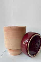 Photo of a Hoff Pot and saucer on a white table in a white room. The pot is rosa clay and sits next to a burgundy glazed saucer.