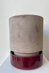 Photo of a Hoff Pot and saucer on a white table in a white room. The pot is gray and sits within to a deep burgundy glazed saucer.