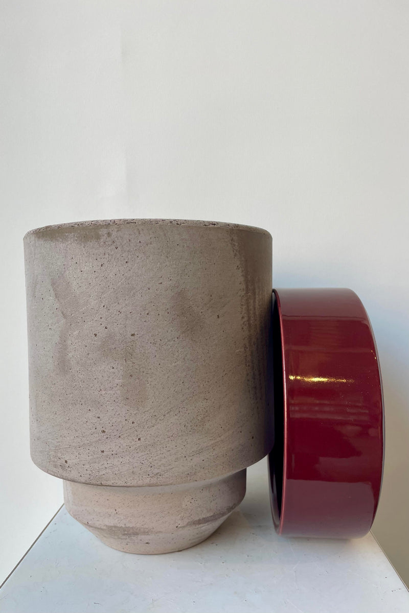 Photo of a Hoff Pot and saucer on a white table in a white room. The pot is gray and sits next to a deep burgundy glazed saucer.