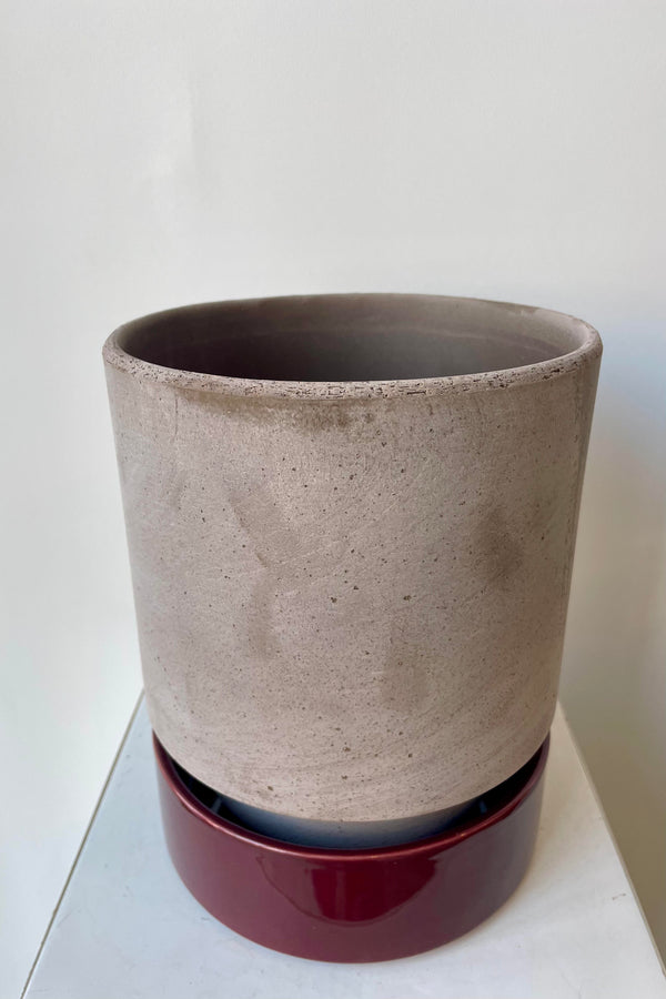 Photo from above of a Hoff Pot and saucer on a white table in a white room. The pot is gray and sits within to a deep burgundy glazed saucer.