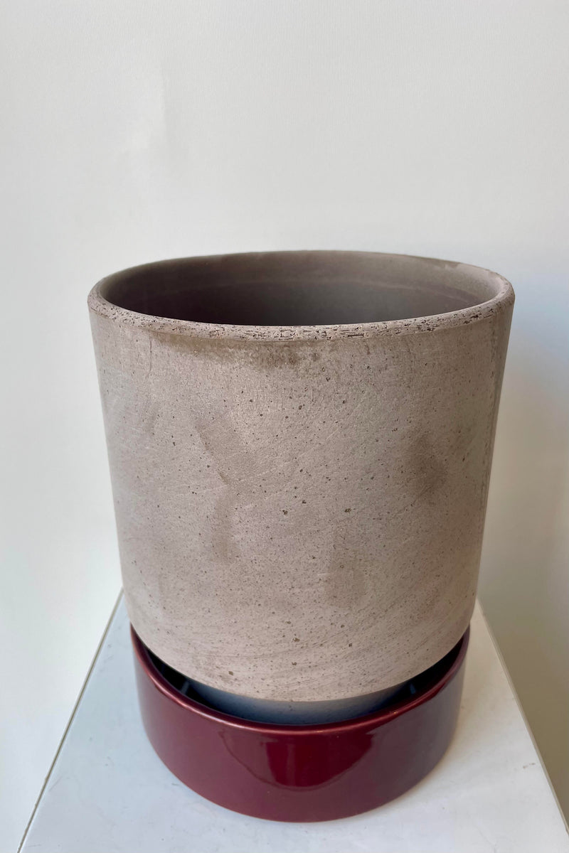 Photo from above of a Hoff Pot and saucer on a white table in a white room. The pot is gray and sits within to a deep burgundy glazed saucer.