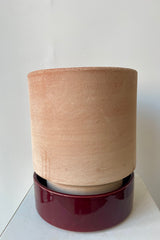 Photo of a Hoff Pot and saucer on a white table in a white room. The pot is rosa clay and sits within a deep burgundy glazed saucer.