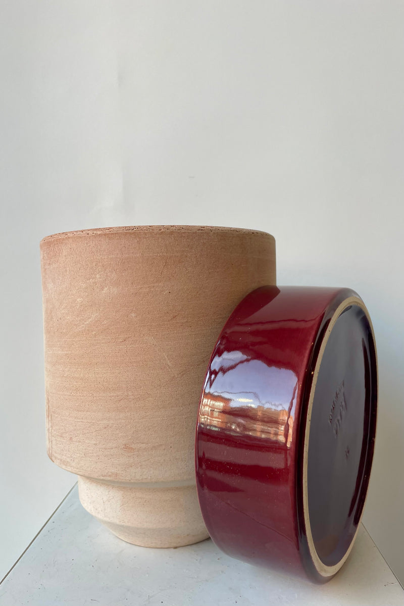 Photo of a Hoff Pot and saucer on a white table in a white room. The pot is rosa clay and sits next to a burgundy glazed saucer.