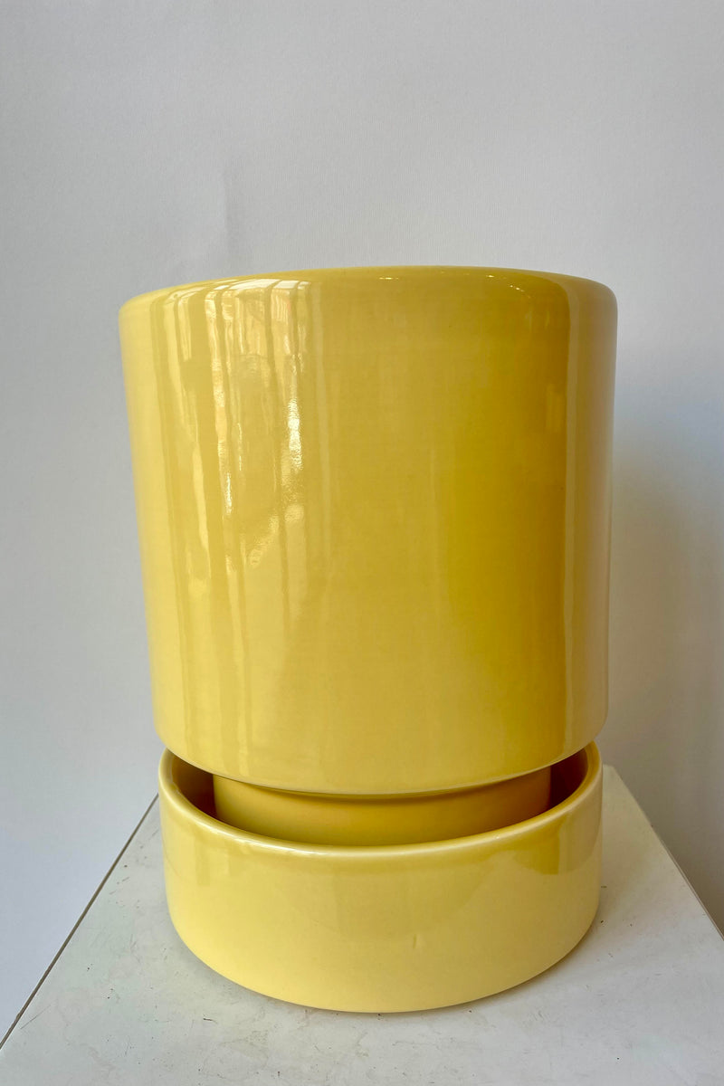 Photo of a Hoff Pot and Saucer set. The glaze of the pottery has a slight sheen and is a pale yellow in color. The pot sits within the deep saucer and the pair are photographed on a white surface in a white room.