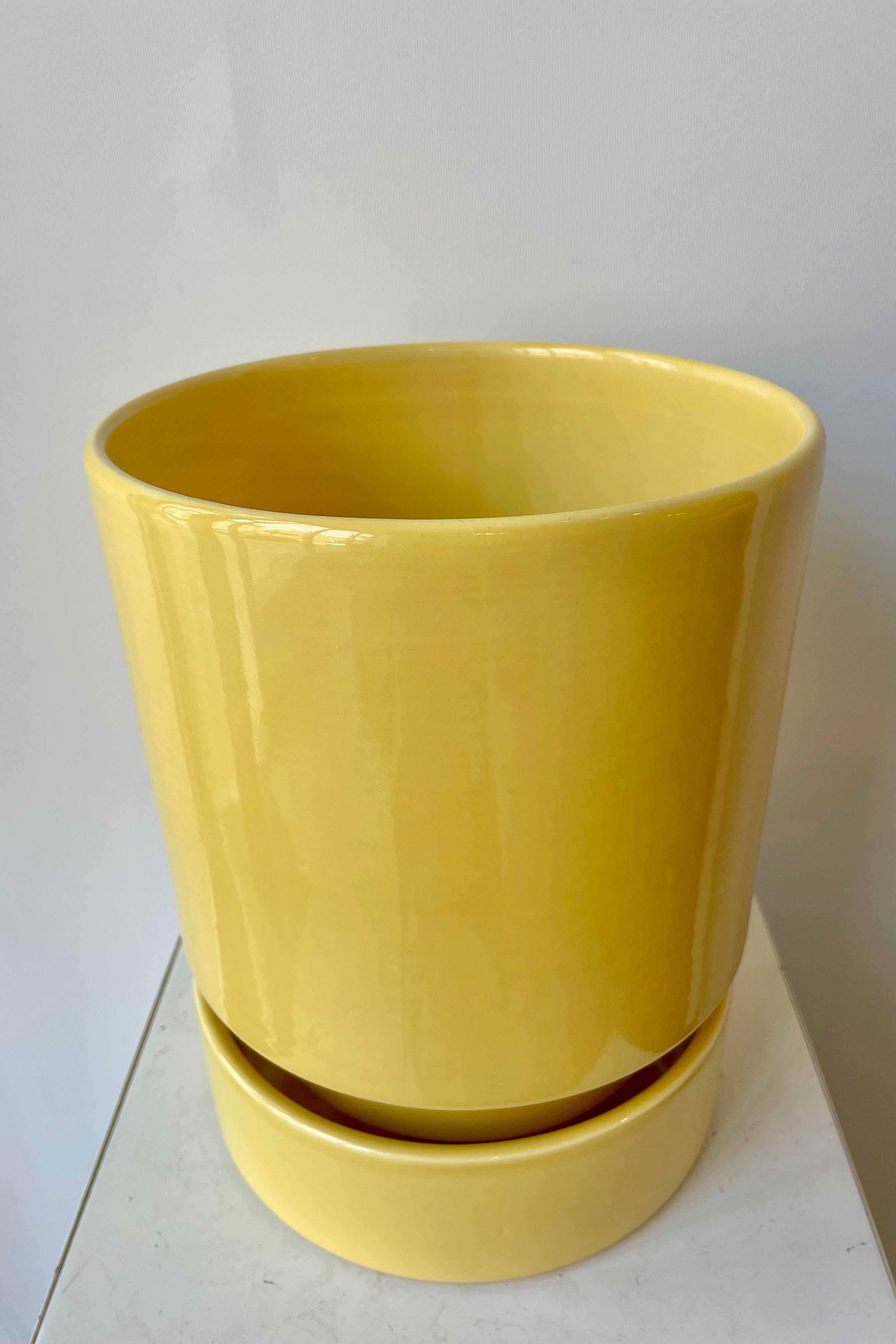 Photo from slightly above of a Hoff Pot and Saucer set. The glaze of the pottery has a slight sheen and is a pale yellow in color. The pot sits within the deep saucer and the pair are photographed on a white surface in a white room.