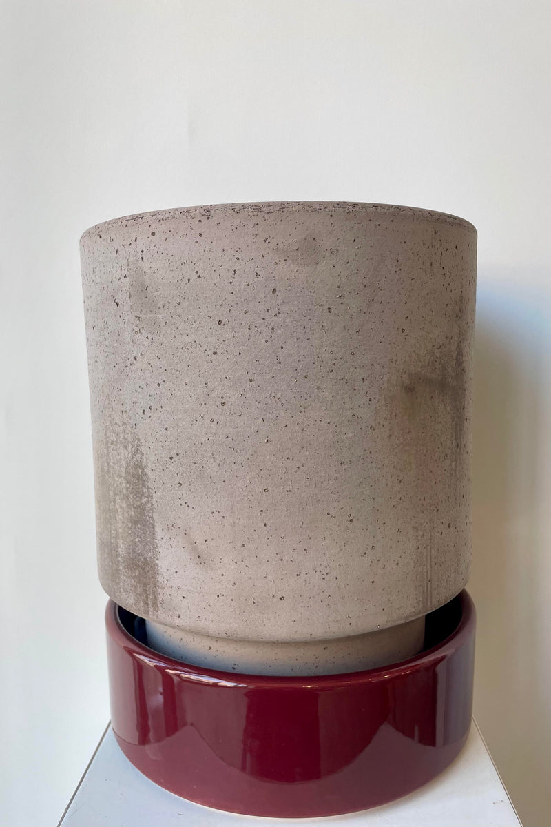 Photo of a Hoff Pot and saucer on a white table in a white room. The pot is gray and sits within a deep burgundy glazed saucer.