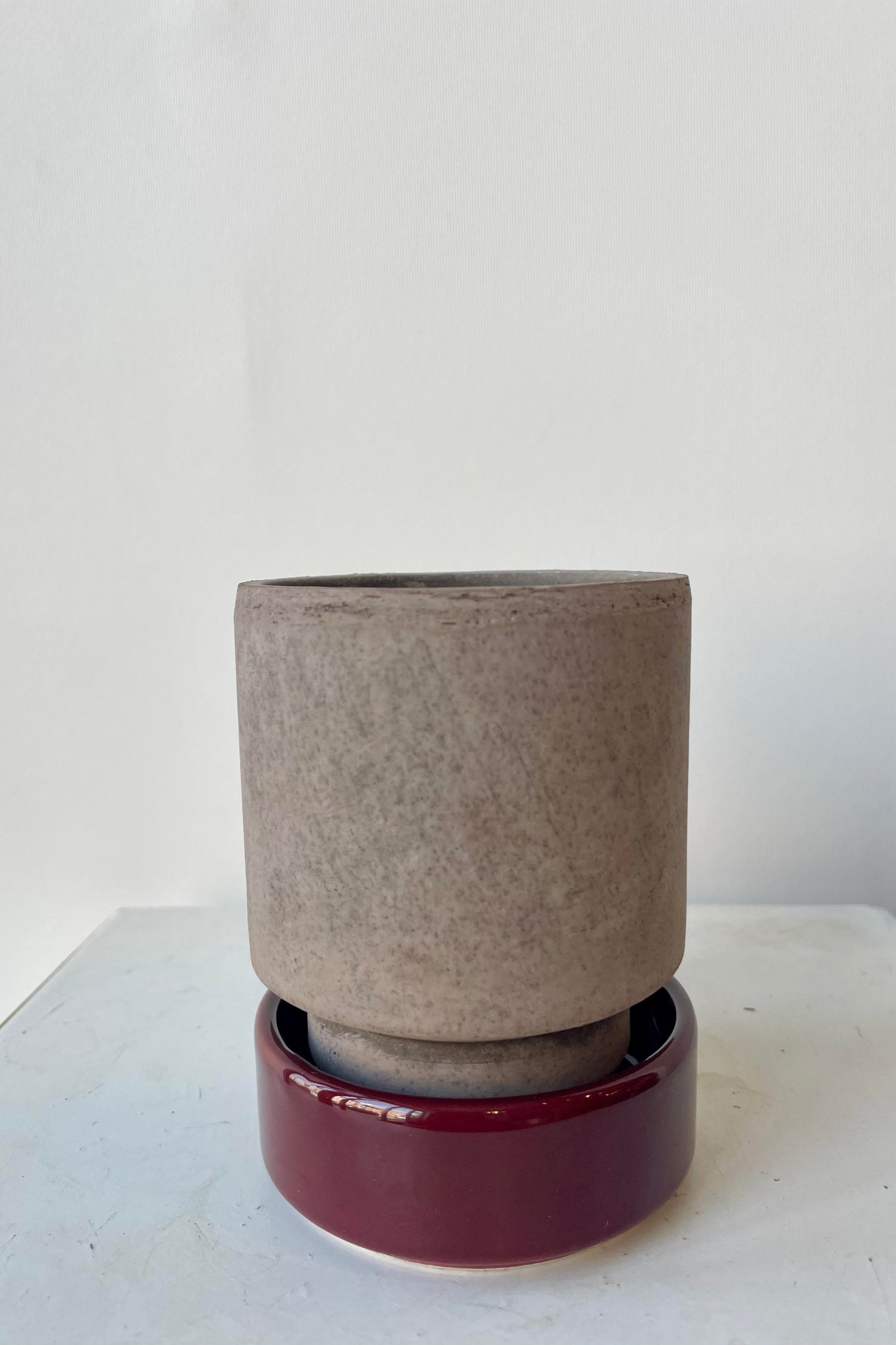Photo of a Hoff Pot and saucer on a white table in a white room. The pot is gray and sits within a deep burgundy glazed saucer. ©Sprout Home