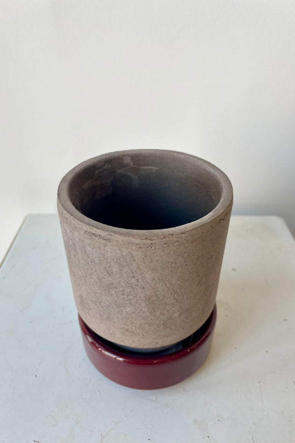 Photo from above of a Hoff Pot and saucer on a white table in a white room. The pot is gray and sits within a deep burgundy glazed saucer.