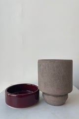 Photo of a Hoff Pot and saucer on a white table in a white room. The pot is gray and sits next to a deep burgundy glazed saucer.