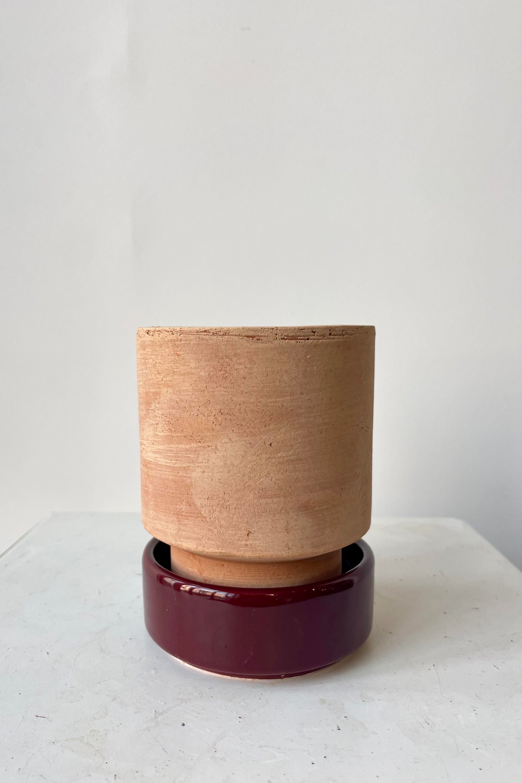 Photo of a Hoff Pot and saucer on a white table in a white room. The pot is rosa unglazed clay and sits within a deep burgundy glazed saucer. ©Sprout Home