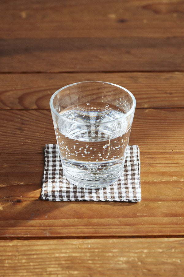 The Joshu Coaster by Fog Linen Work sitting on a wood table with a glass of water on top of it. 