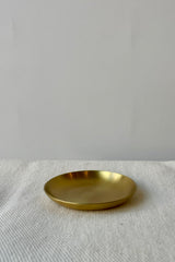 Photo of an extra extra small brass plate with a 2" diameter on a white cloth against a white wall. Shown from the side.