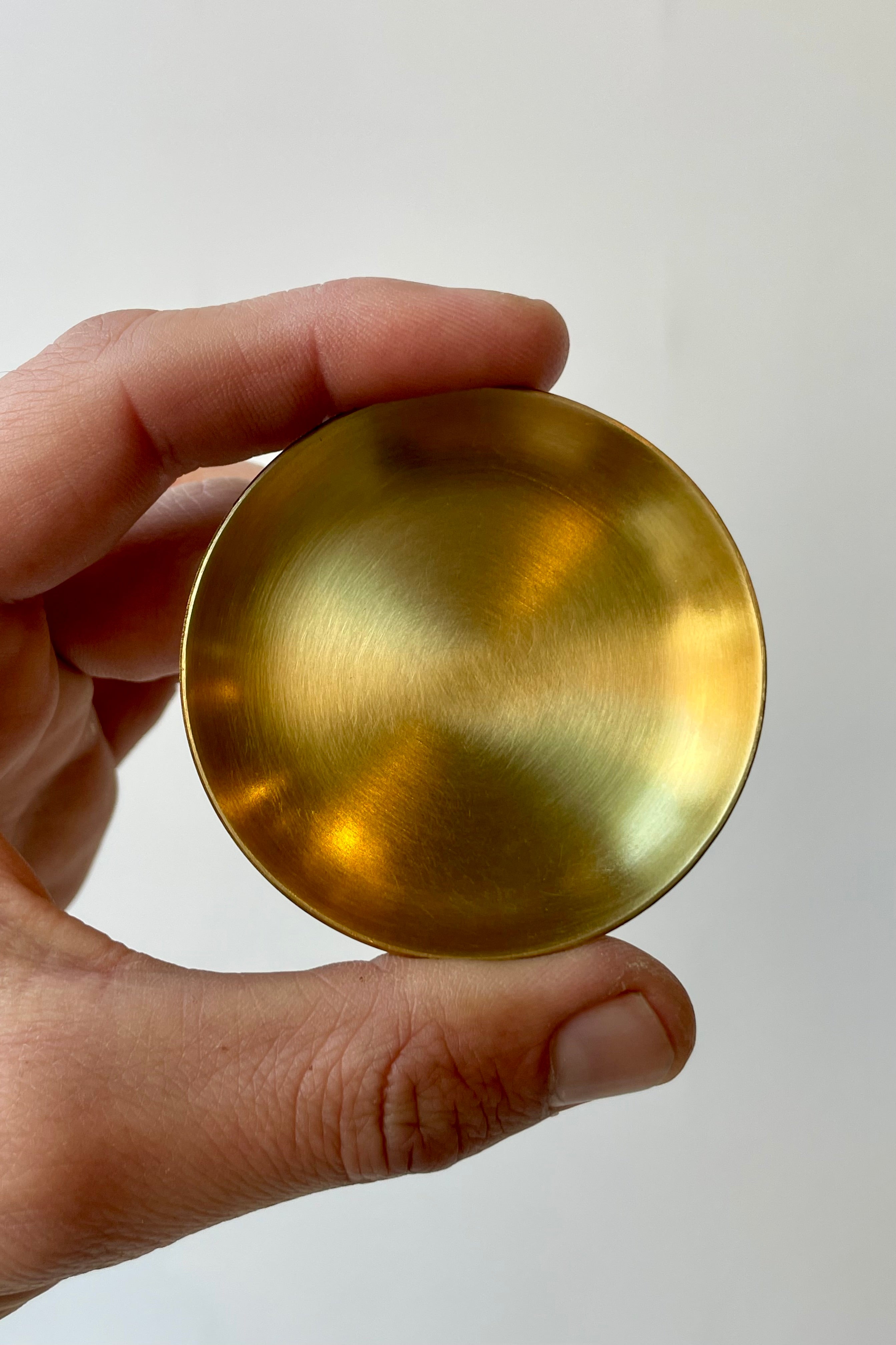 Photo of an extra extra small brass plate with a 2" diameter against a white wall. A hand is holding the plate on its side between the thumb and forefinger.