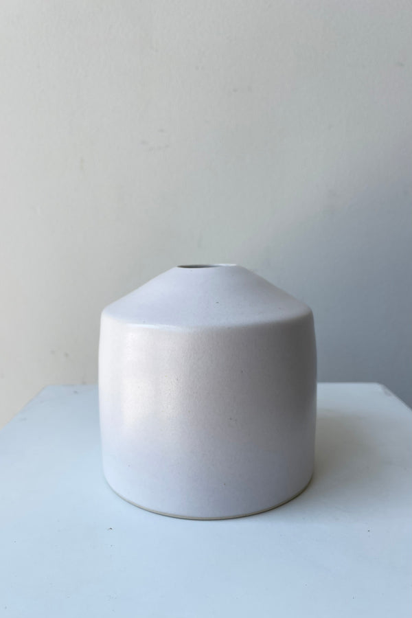 One small ceramic vase sits on a white surface in a white room. The vase is cylindrical and tapers off at the top to a narrow opening. The vase is white with a small ring of unglazed clay at the bottom. It is photographed straight on.