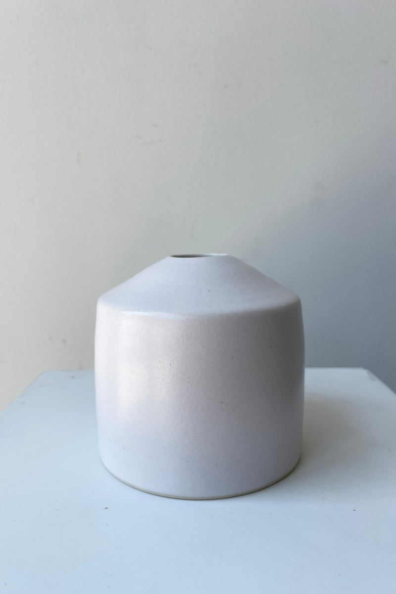 One small ceramic vase sits on a white surface in a white room. The vase is cylindrical and tapers off at the top to a narrow opening. The vase is white with a small ring of unglazed clay at the bottom. It is photographed straight on.