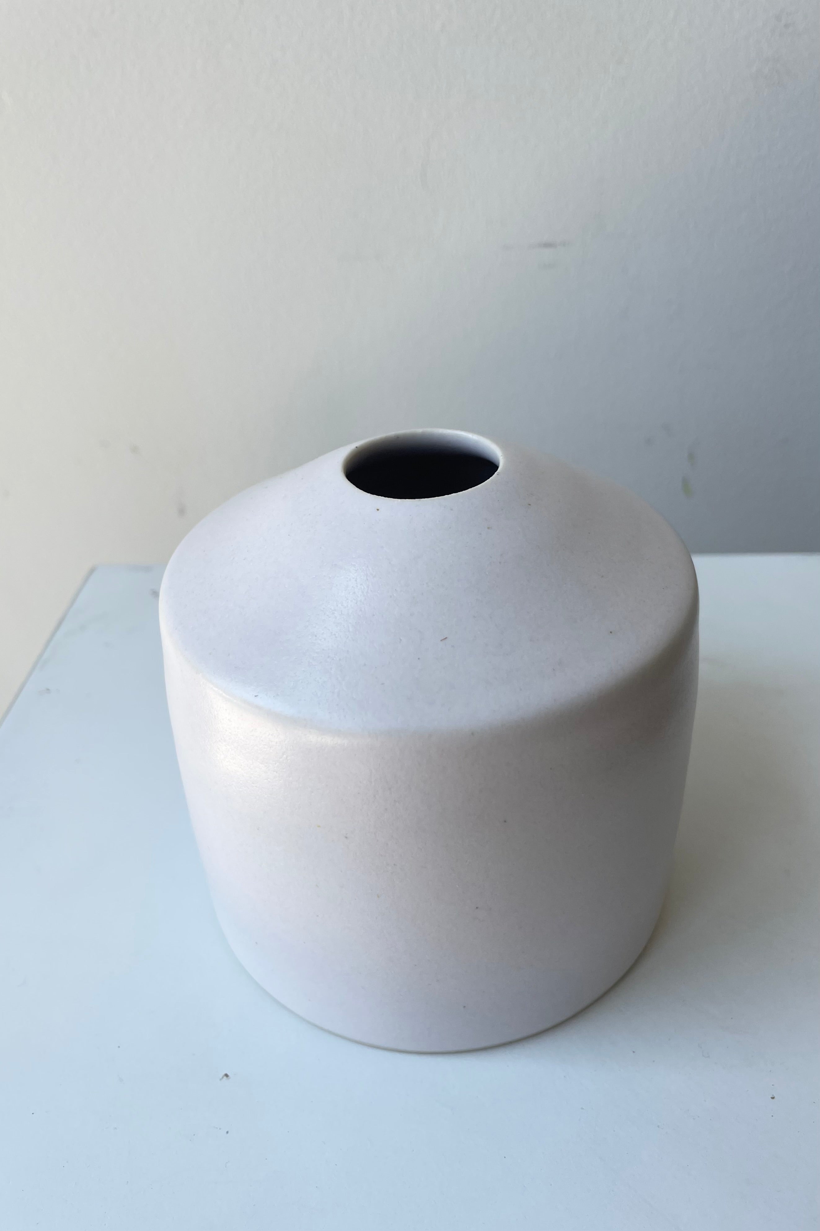 One small ceramic vase sits on a white surface in a white room. The vase is cylindrical and tapers off at the top to a narrow opening. The vase is white with a small ring of unglazed clay at the bottom. It is photographed from an above angle.