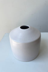 One small ceramic vase sits on a white surface in a white room. The vase is cylindrical and tapers off at the top to a narrow opening. The vase is white with a small ring of unglazed clay at the bottom. It is photographed from an above angle.