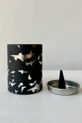 A black cardboard cylinder featuring silver patterning and type with a circular silver tray and one cone incense on a white table with white background. This scent is called Death and the fragrance contains notes of leather, marble and sage.