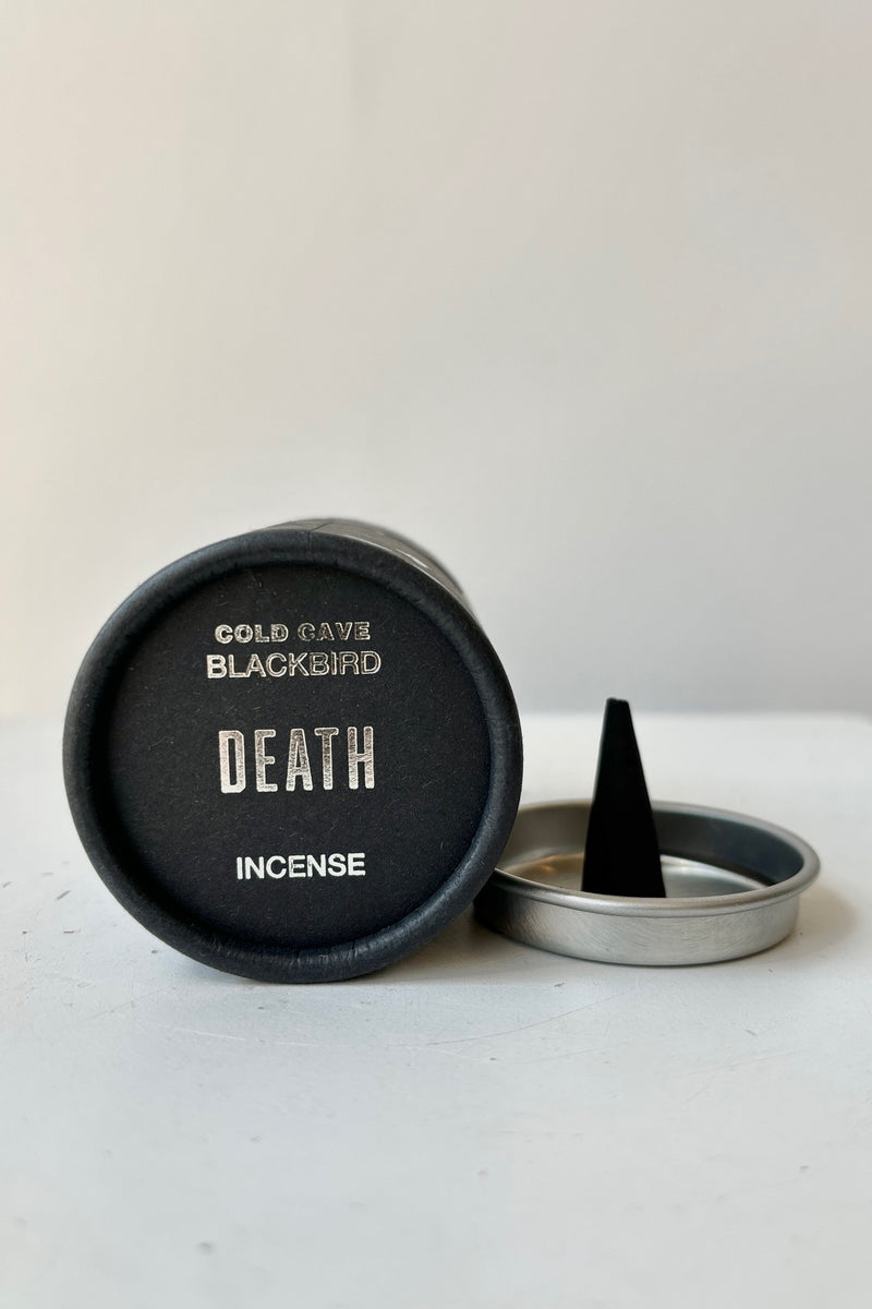 A black cardboard cylinder featuring silver patterning and type with a circular silver tray and one cone incense on a white table with white background. This scent is called Death and the fragrance contains notes of leather, marble and sage.