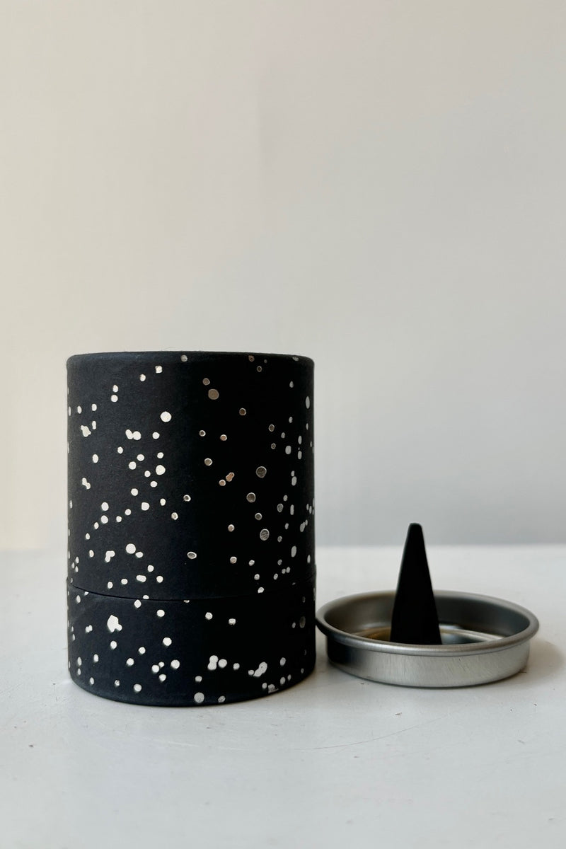 A black cardboard cylinder featuring silver patterning and type with a circular silver tray and one cone incense on a white table with white background. This scent is called Love and the fragrance contains notes of honey, rose and ivy.