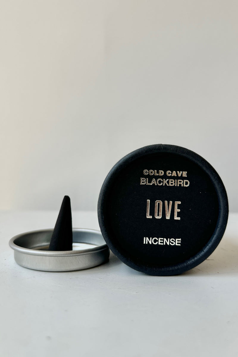 A black cardboard cylinder featuring silver patterning and type with a circular silver tray and one cone incense on a white table with white background. This scent is called Love and the fragrance contains notes of honey, rose and ivy.