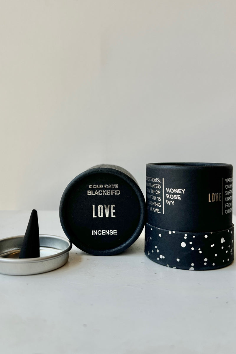 A black cardboard cylinder featuring silver patterning and type with a circular silver tray and one cone incense on a white table with white background. This scent is called Love and the fragrance contains notes of honey, rose and ivy.