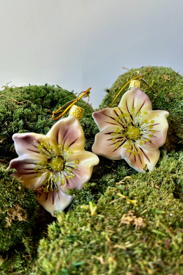 Two white glass ornaments with splashes of purple on the petals with pale green glitter displayed on a moss mound against a white wall. 