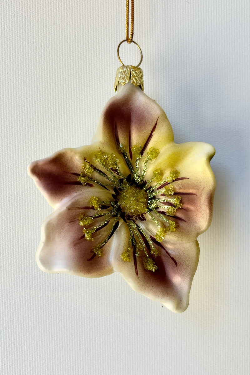 A white glass ornament in the shape of a Helleborus flower with splashes of purple on the petals with pale green glitter against a white background.