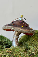 The purple wooded glen mushroom ornament has a brown frosted cap accented with multicolored gems and glitter throughout displayed on bed of moss against a white wall. 