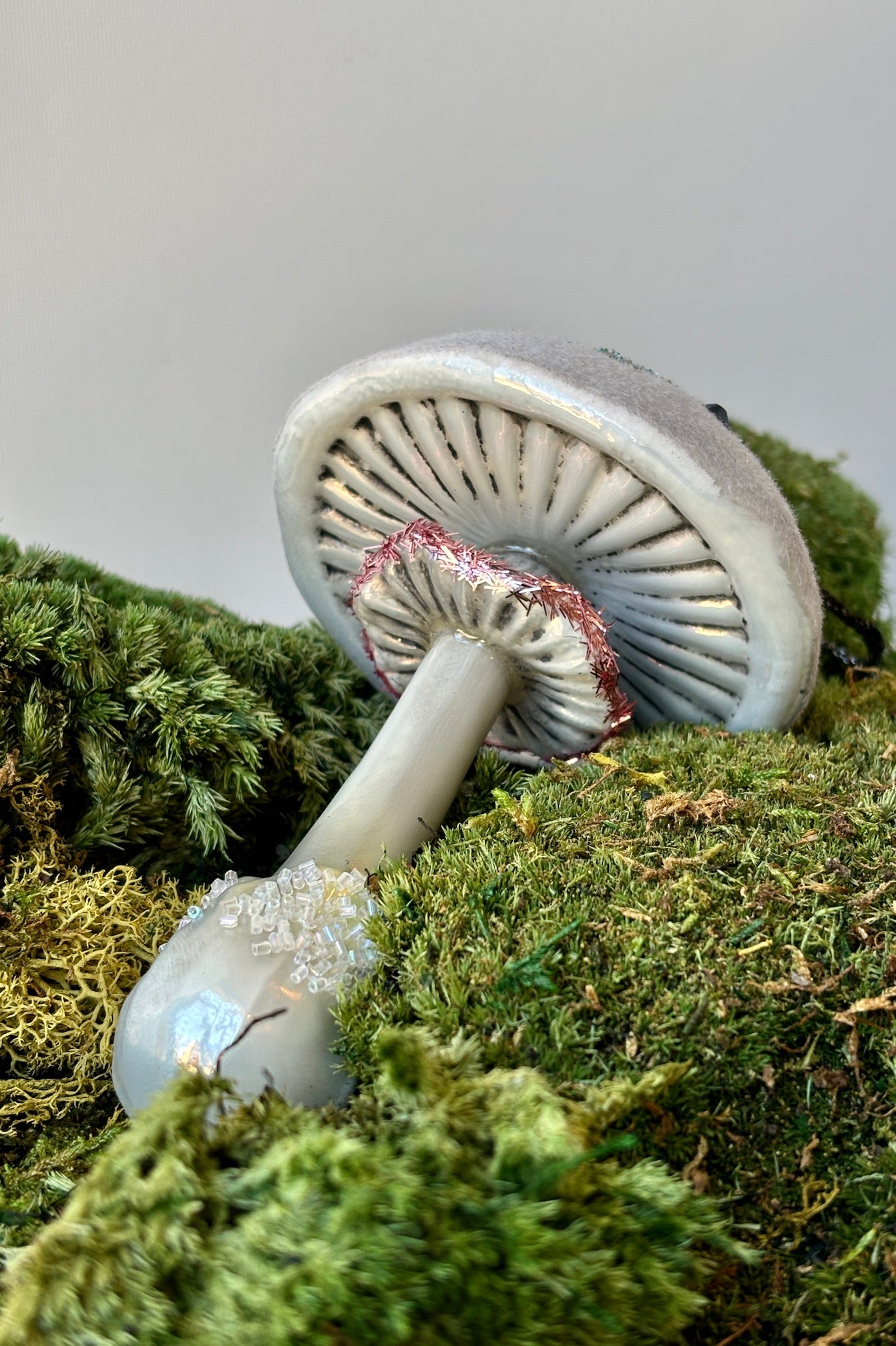 Underside of Frostfield mushroom in white glass with an opalescent glitter frosted cap accented with blue gemstones and a small ruffled underneath outlined in pink shredded glitter on a mound of moss.