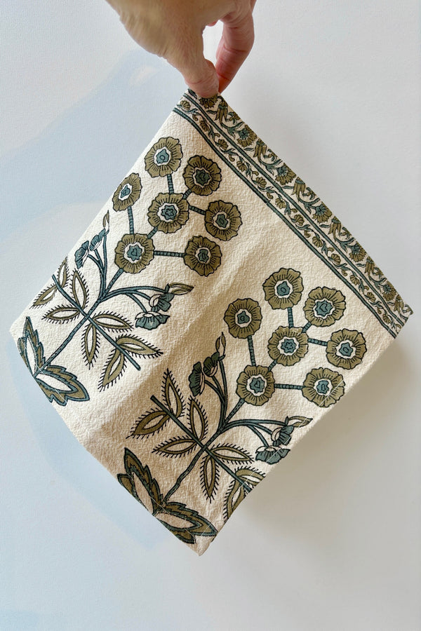 One hundred percent cotton napkin with a hand block printed floral motif in teal and gold with a floral printed border against a white wall.