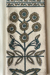 Detail of a one hundred percent cotton napkin with a hand block printed floral motif in teal and gold with a floral printed border against white background.