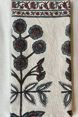 Detail of one hundred percent cotton napkin with a hand block printed floral motif in teal and ruby with a floral printed border against a white wall.