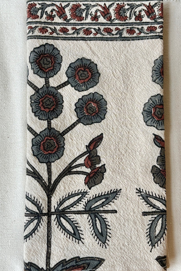 Detail of one hundred percent cotton napkin with a hand block printed floral motif in teal and ruby with a floral printed border against a white wall.