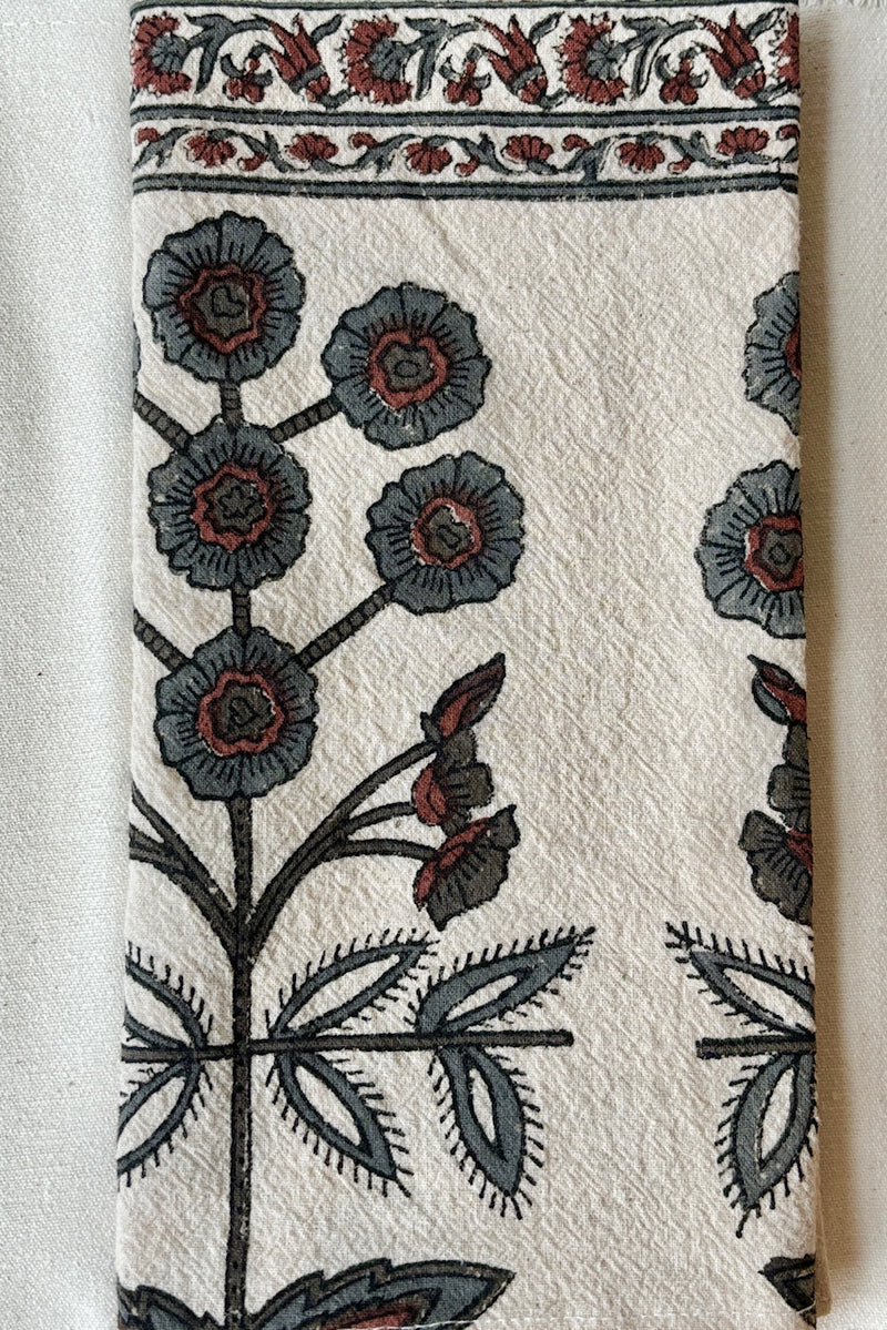 Detail of one hundred percent cotton napkin with a hand block printed floral motif in teal and ruby with a floral printed border against a white wall.