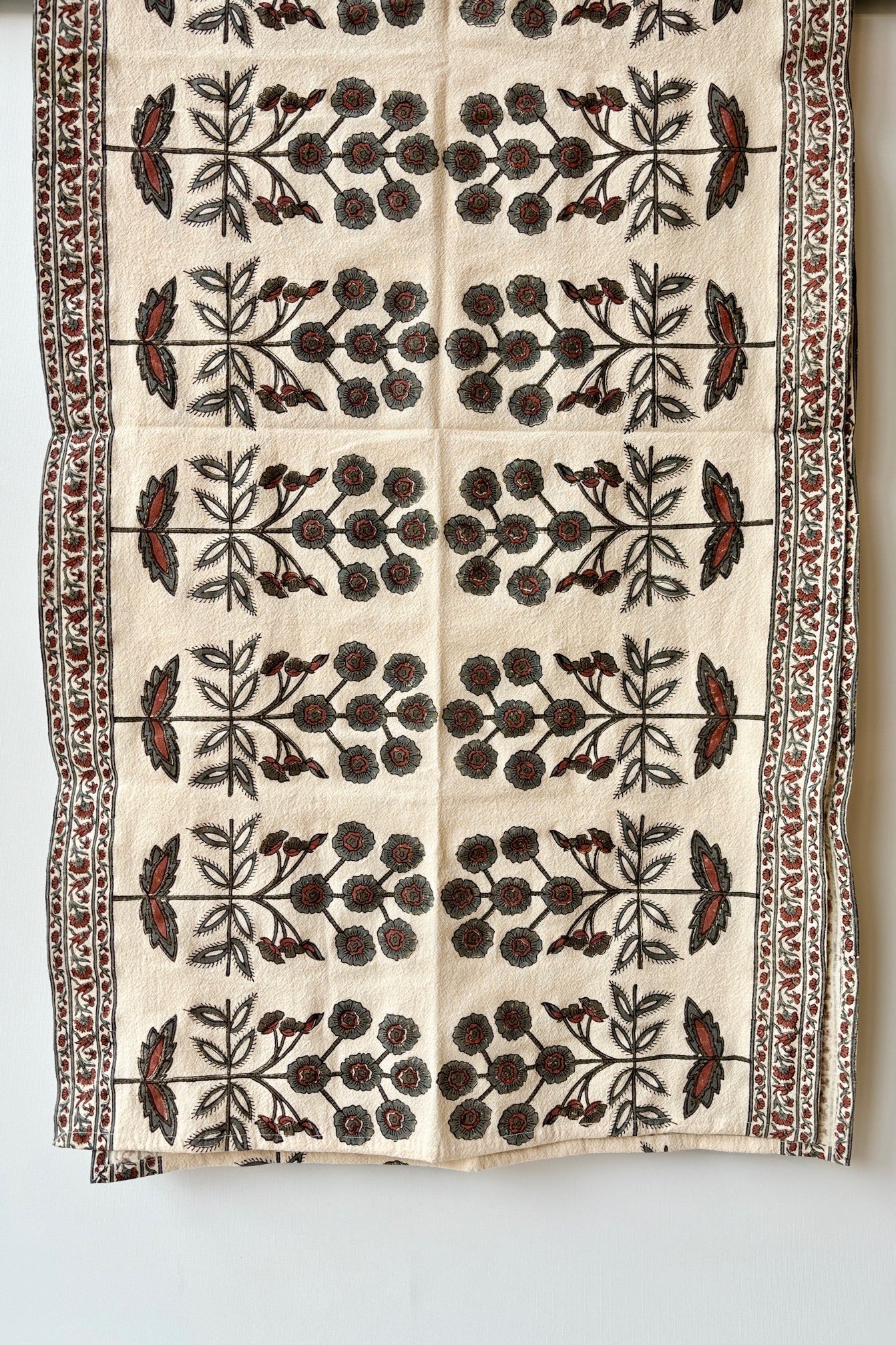 One hundred percent cotton table runner with a teal and ruby floral motif arranged in a symmetrical pattern with a border of smaller floral patterns at the edges. 