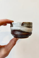 Hand golding the Bizan sake cup with clay body and brown, white and gray glazing against white background.