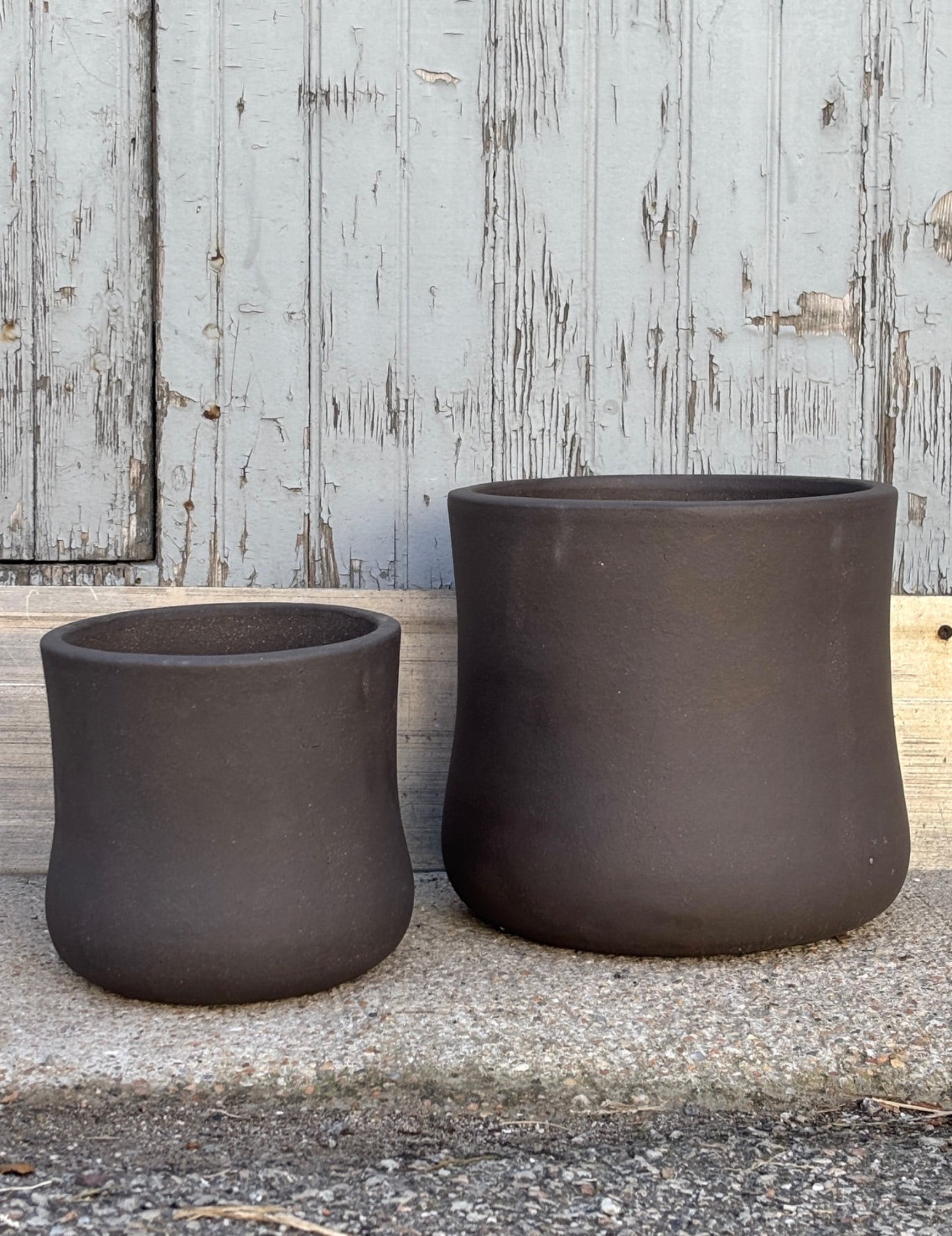 Medium  and small Brown Terra cotta pots with hourglass shape against a grey wall on a cement surface. Planters contain a drainage hole. 