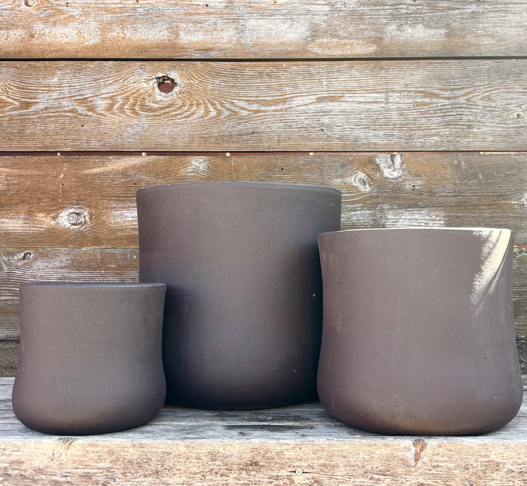 Trio of Brown Terra cotta pots with an hourglass shape against a wood wall. Planters contain a drainage hole. 