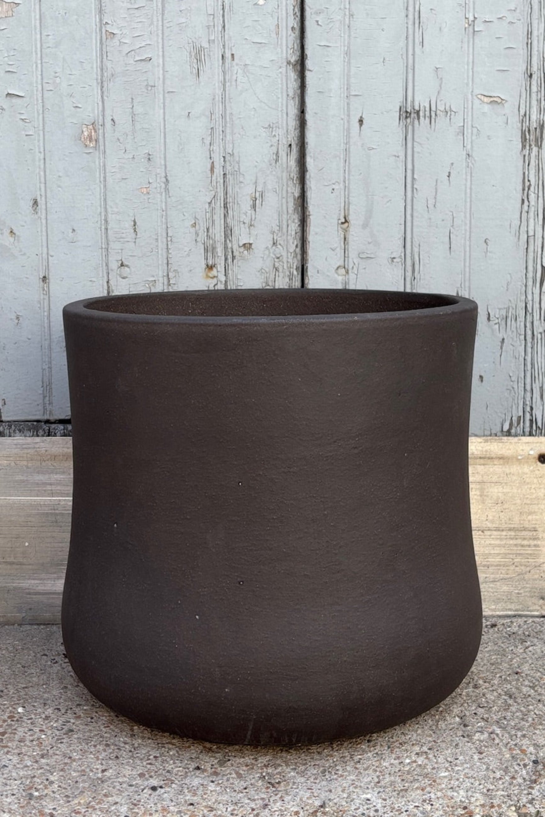 Large Brown Terra cotta pot with an hourglass shape against a grey wall on a cement surface. Planter contains a drainage hole. 