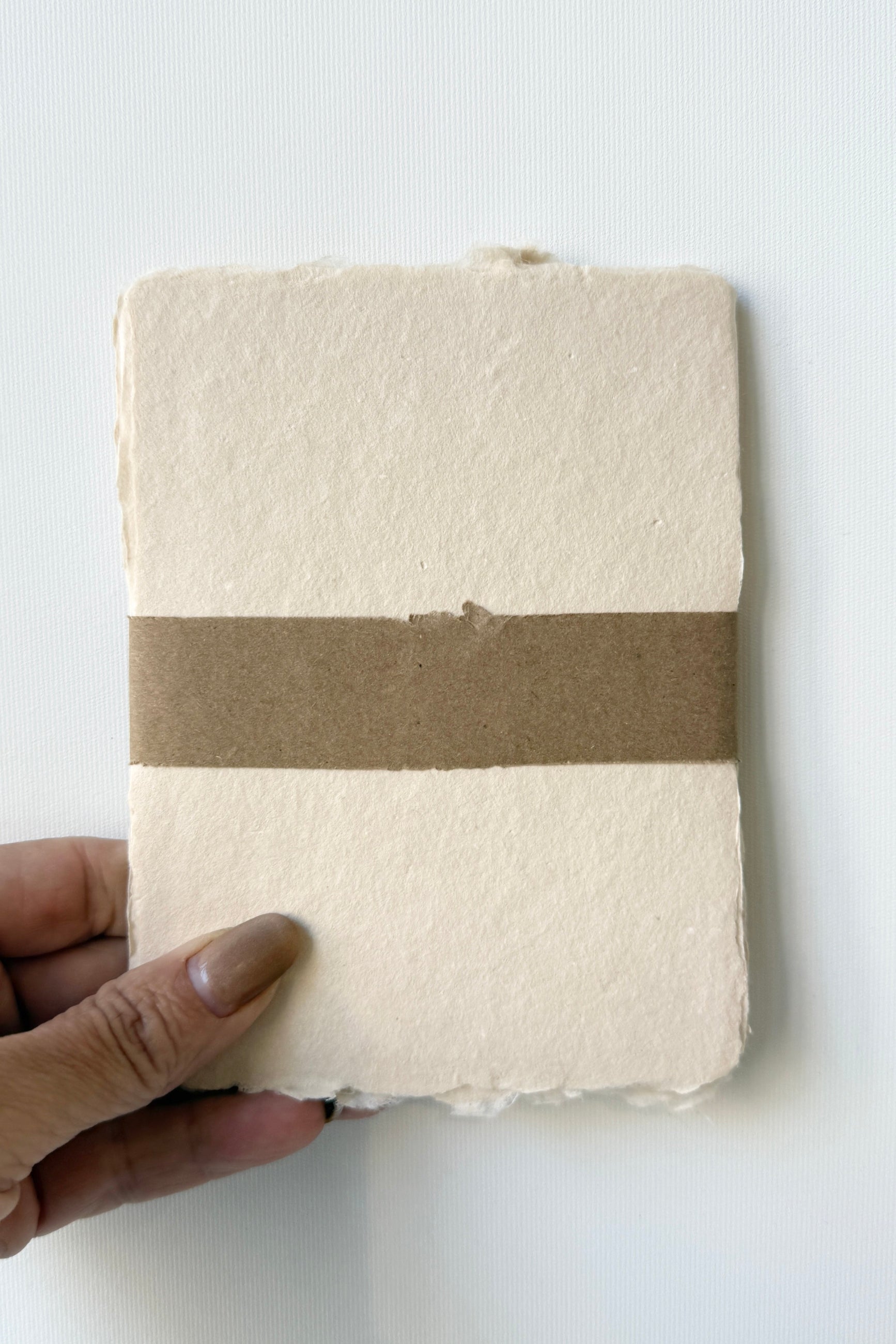 Hand holding a Handmade Buff Paper Sheet Set in A2 with a brown band by Sustainable Paper + Craft against a white background ©Sprout Home
