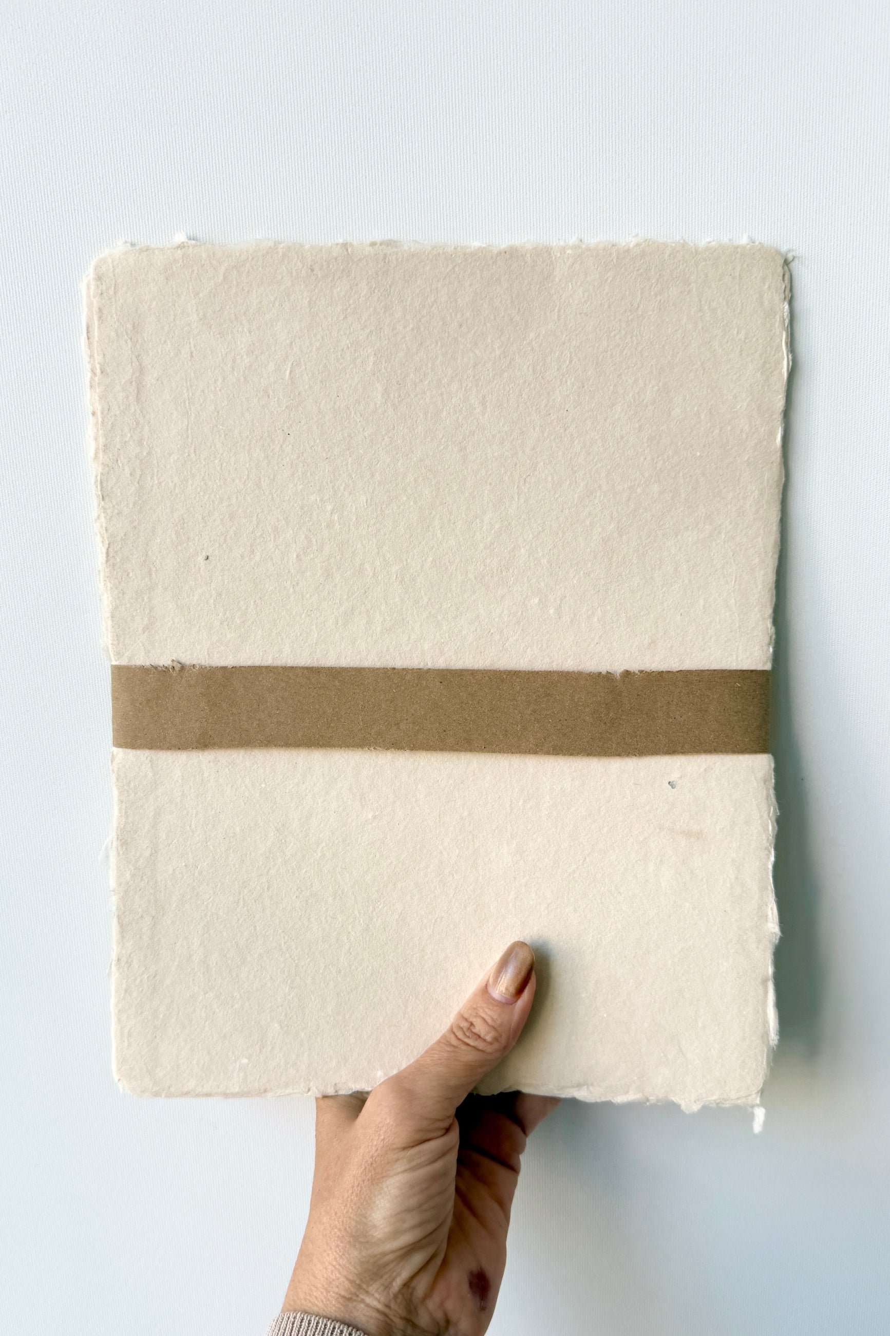 Hand holding a fHandmade Buff Paper Sheet Set in 8x10 with a brown band by Sustainable Paper & Craft against a light gray background ©Sprout Home